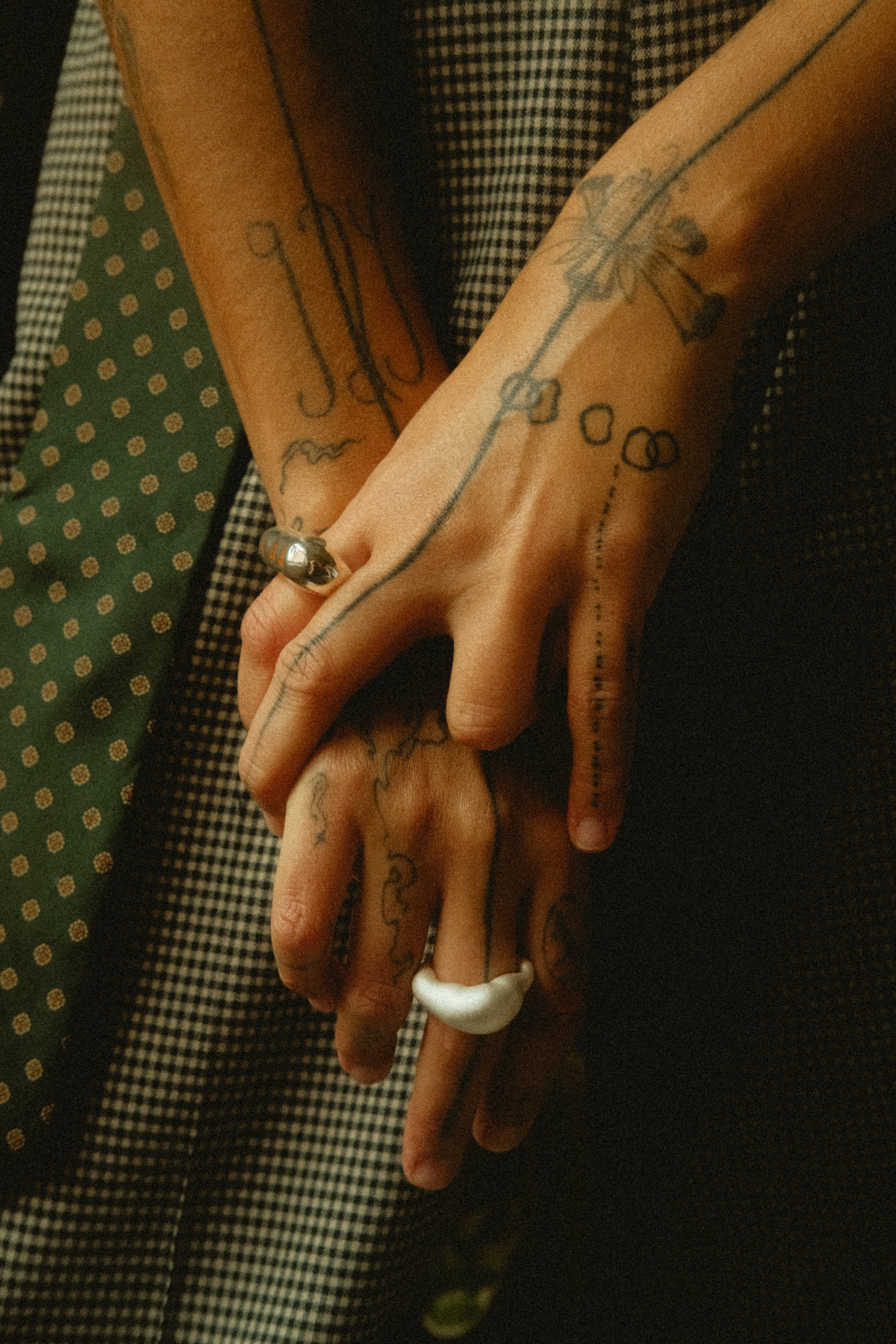 Close-up of hands with tattoos holding each other, wearing rings, against a dark background.